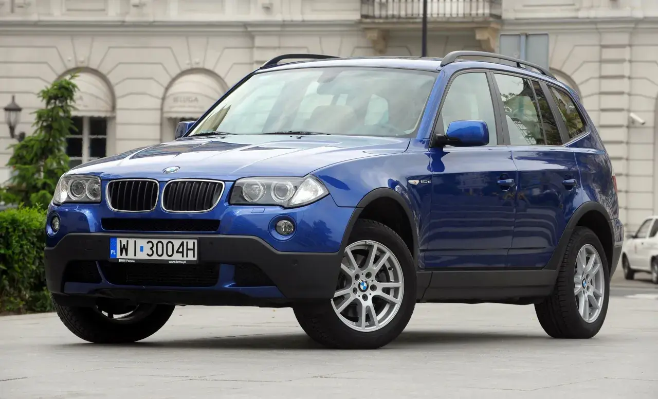 Niebieskie BMW X3 2005 z polskimi tablicami rejestracyjnymi WI 3004H zaparkowane przed budynkiem.