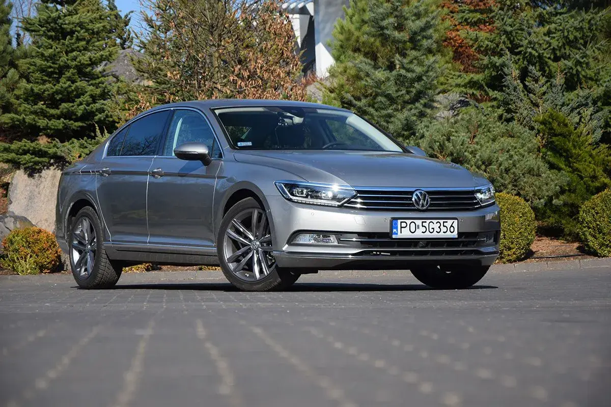 Szary Volkswagen Passat sedan zaparkowany na podjeździe, otoczony zielenią i kamieniami.