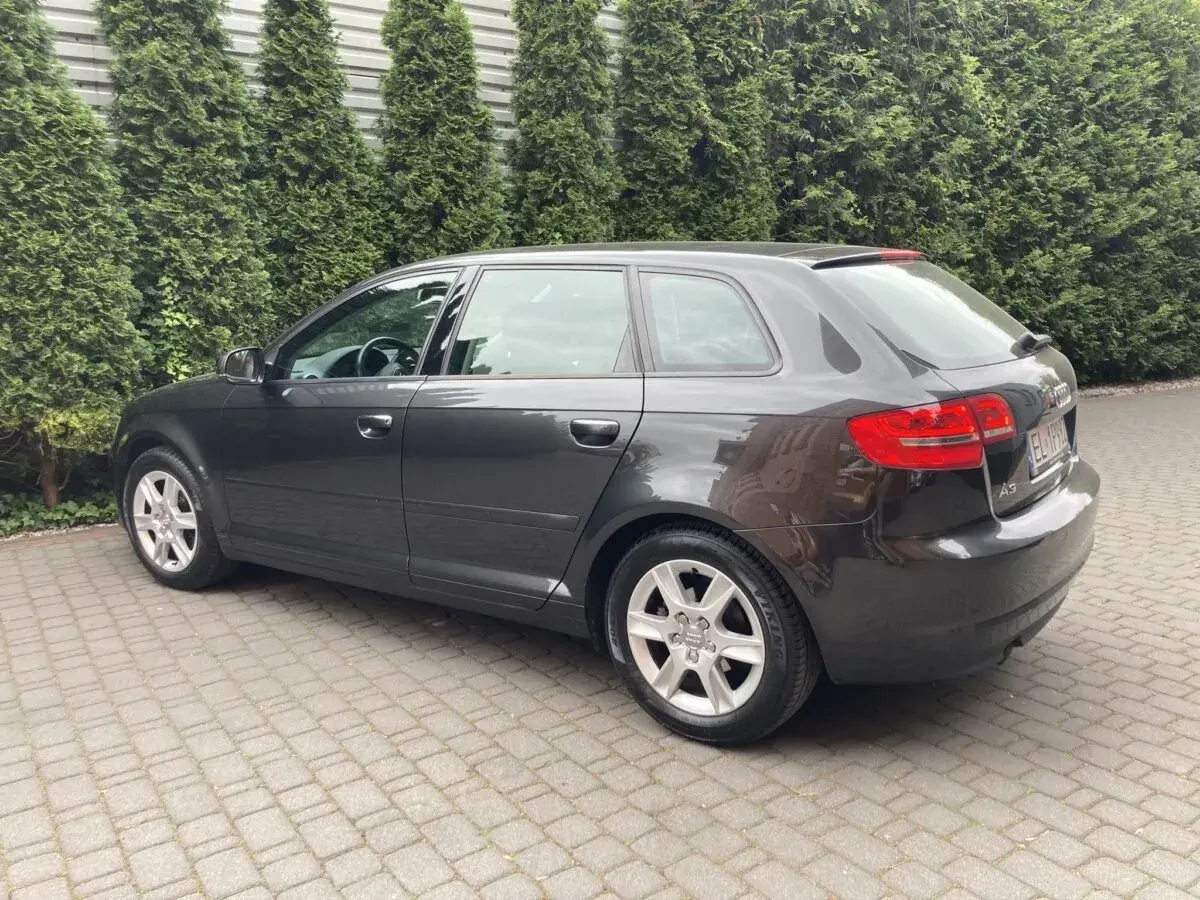Ciemnoszary Audi A3 8P Sportback stoi na podjeździe z kostki brukowej, otoczony zielonym żywopłotem.