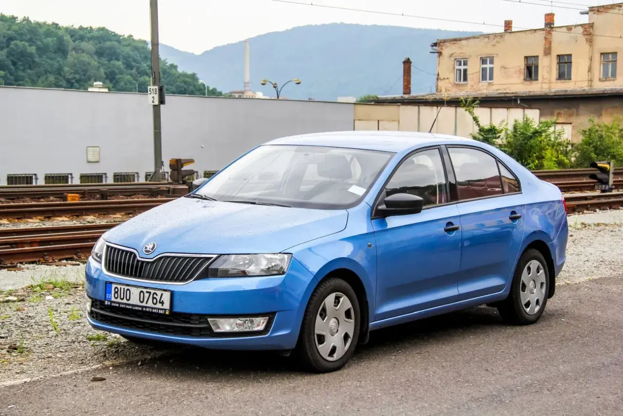 Niebieski liftback Skoda Rapid stoi na tle torów kolejowych i starych budynków.