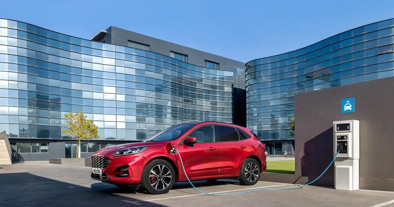 Czerwony Ford Kuga, nowoczesny kombi, ładuje się przy stacji ładowania przed futurystycznym budynkiem.