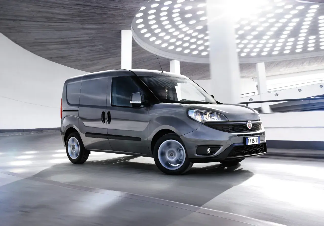Szary Fiat Doblo Cargo w ruchu, z rozmytymi kołami, na tle nowoczesnej architektury z okrągłymi światłami.