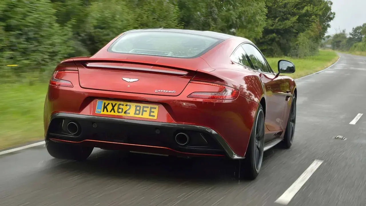 Czerwony Aston Martin Vanquish pędzi po mokrej drodze, otoczony zielenią.