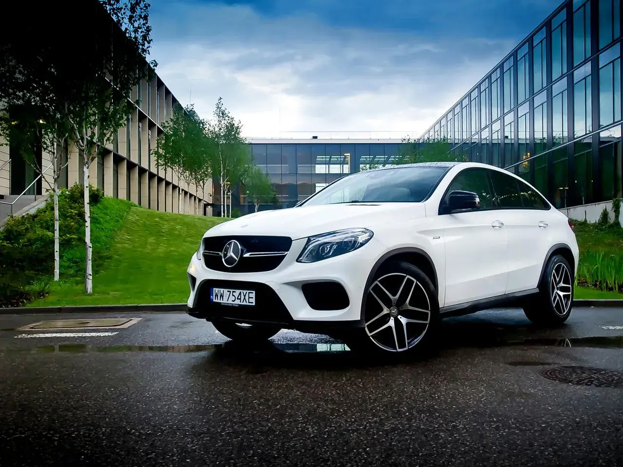 Biały Mercedes GLE 450 zaparkowany na mokrym asfalcie przed nowoczesnym budynkiem.