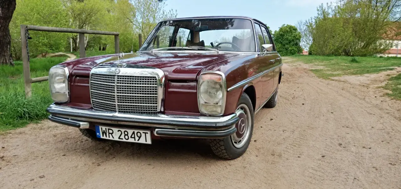 Klasyczny mercedes w114 w bordowym kolorze, zaparkowany na piaszczystej drodze wśród zieleni.