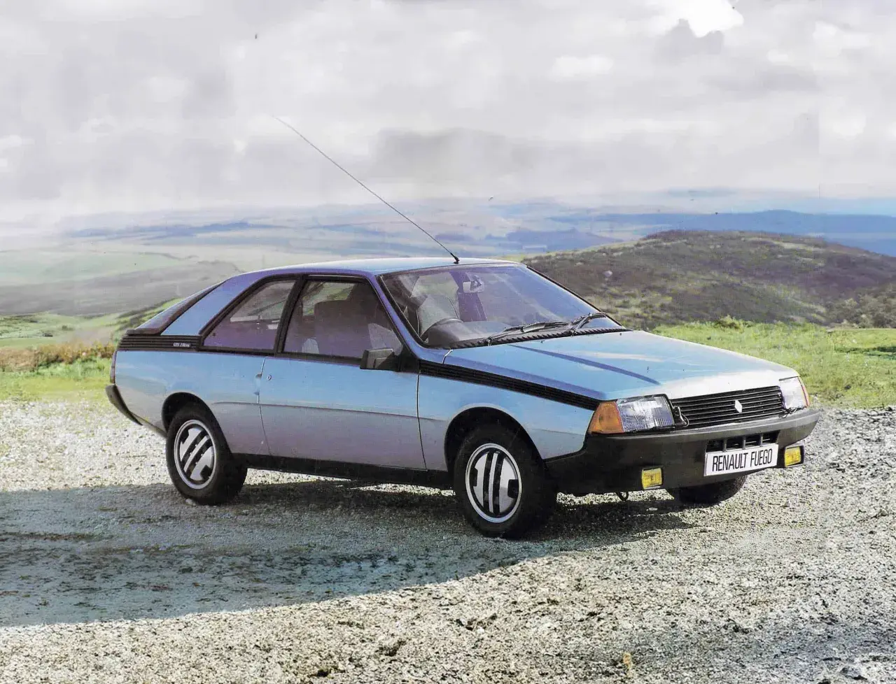 Jasnoniebieski Renault Fuego z lat 80. stoi na szutrowej drodze, z widokiem na pagórkowaty krajobraz i chmurne niebo.