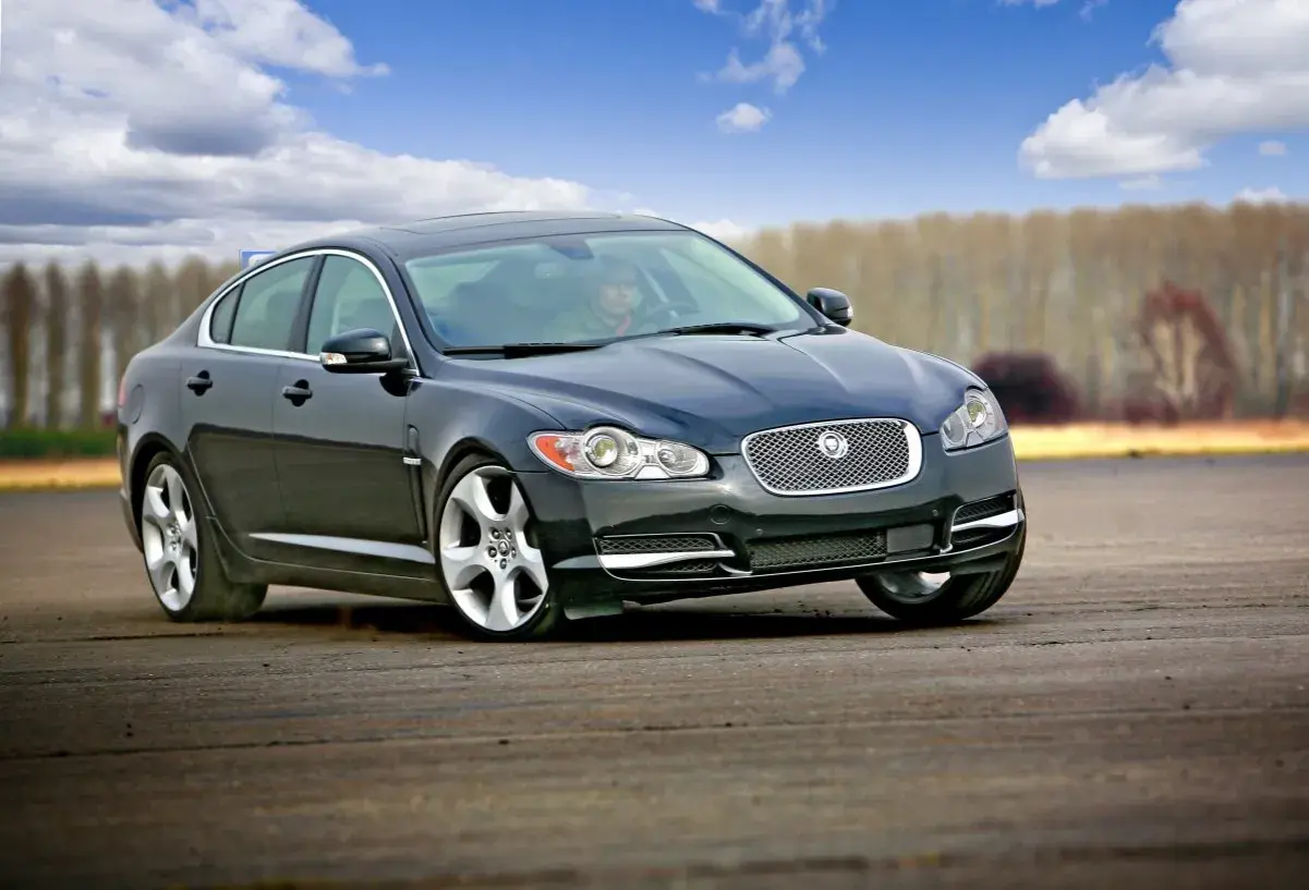 Czarny Jaguar XF na torze. Kierowca w środku, chmury na niebie. Wiele pozytywnych jaguar xf opinie potwierdza jego sportowy charakter.