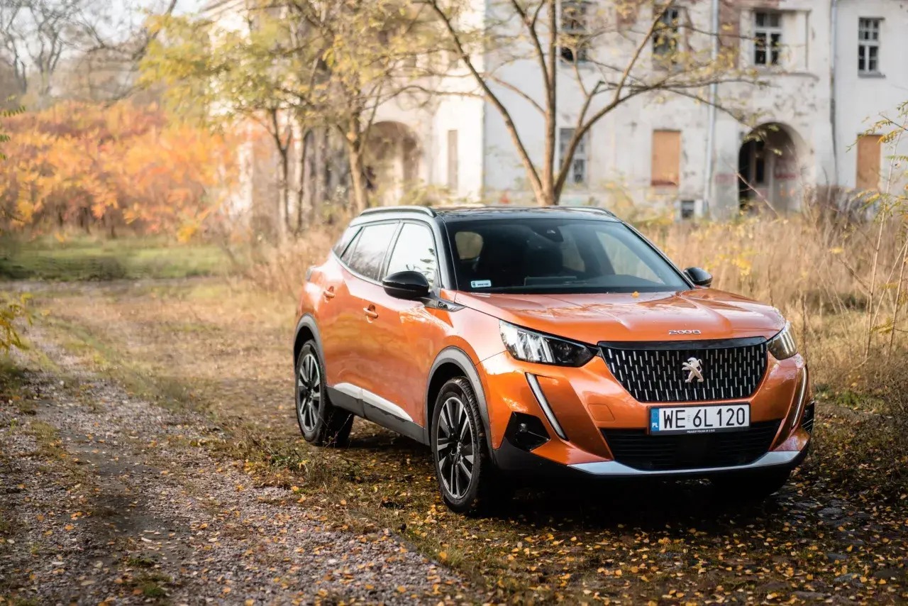 Pomarańczowy Peugeot 2008 GT na tle jesiennego krajobrazu z opuszczonym budynkiem.