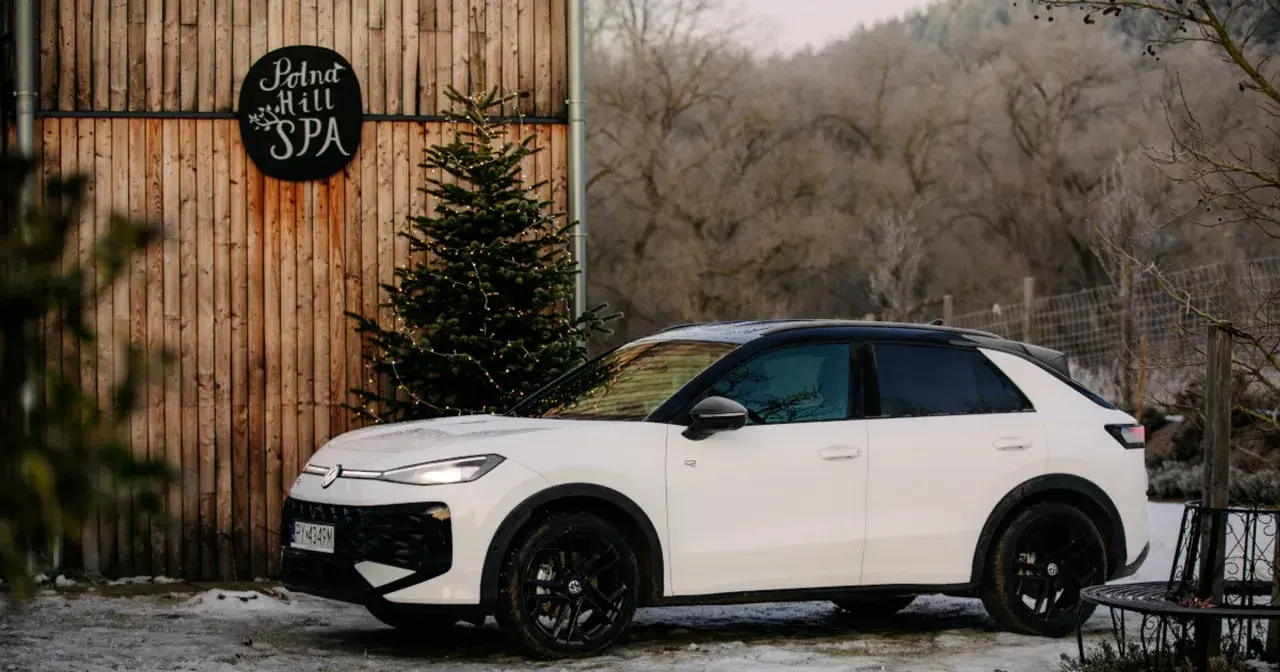 Biały, mały SUV zaparkowany obok drewnianej chaty z napisem "Polna Hill SPA" i choinką ozdobioną lampkami.