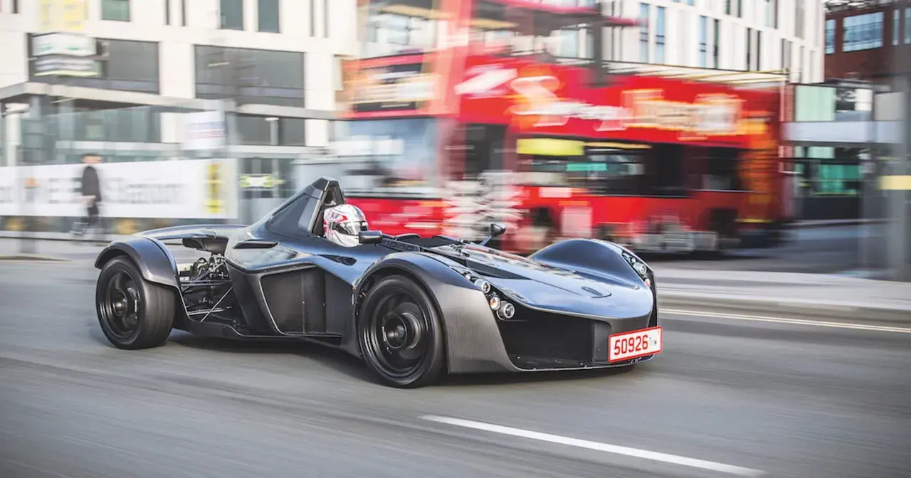 Szary BAC Mono z kierowcą w kasku pędzi ulicą, mijając rozmazany czerwony autobus piętrowy.
