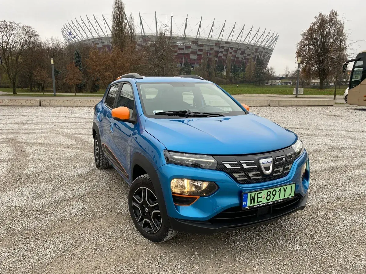 Niebieska Dacia Spring z pomarańczowymi akcentami na tle Stadionu Narodowego. Dacia Spring opinie – miejski elektryk gotowy na przygodę.