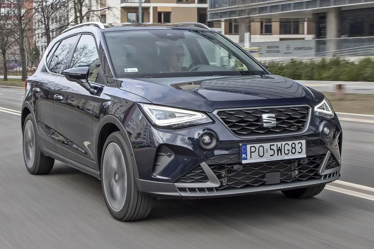 Ciemnoniebieski Seat Arona w ruchu ulicznym. Kierowca w okularach. Wiele pozytywnych seat arona opinie potwierdza jego miejski charakter.