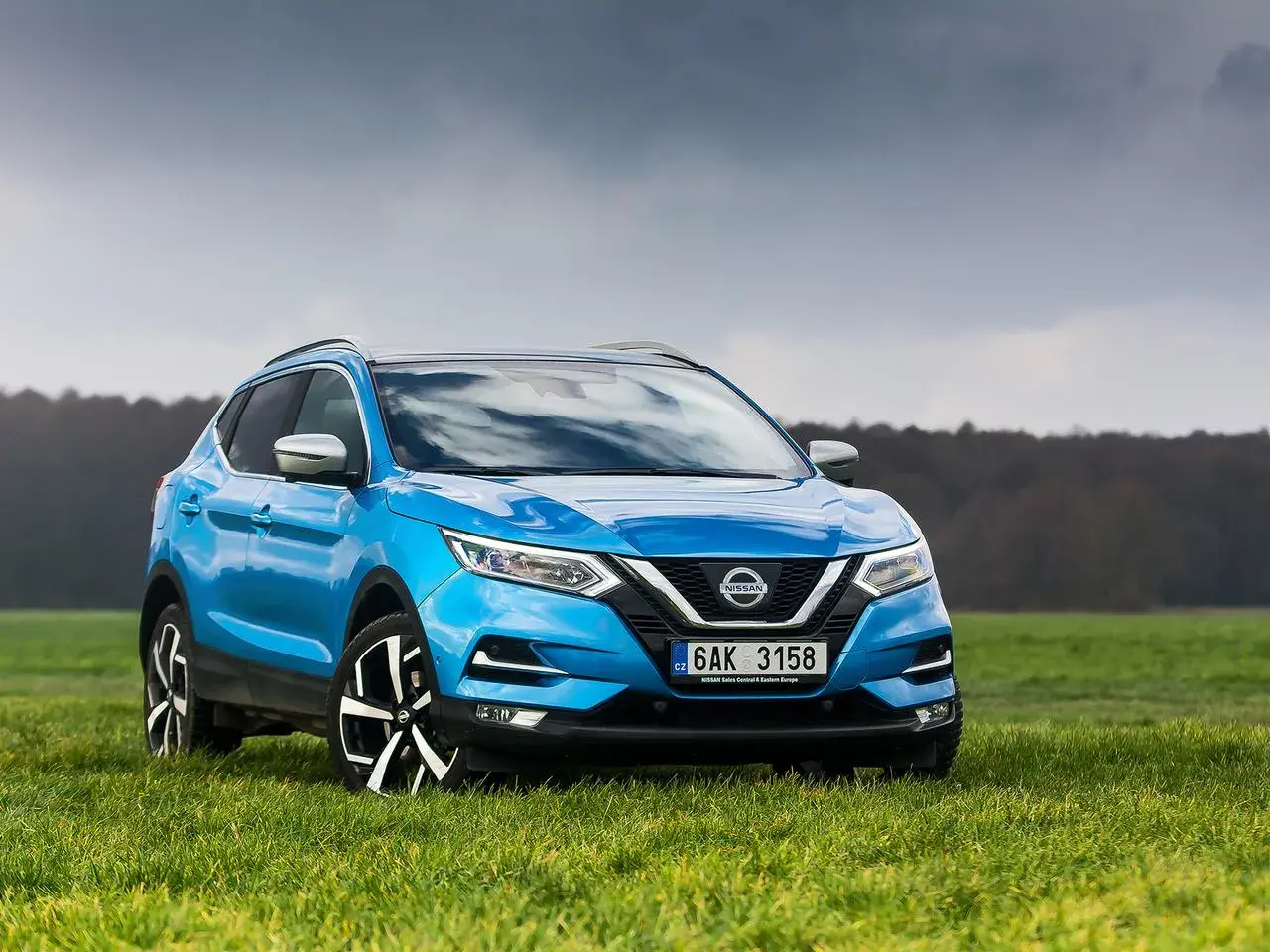 Niebieski Nissan Qashqai 1.6 benzyna na tle zielonej łąki i pochmurnego nieba.
