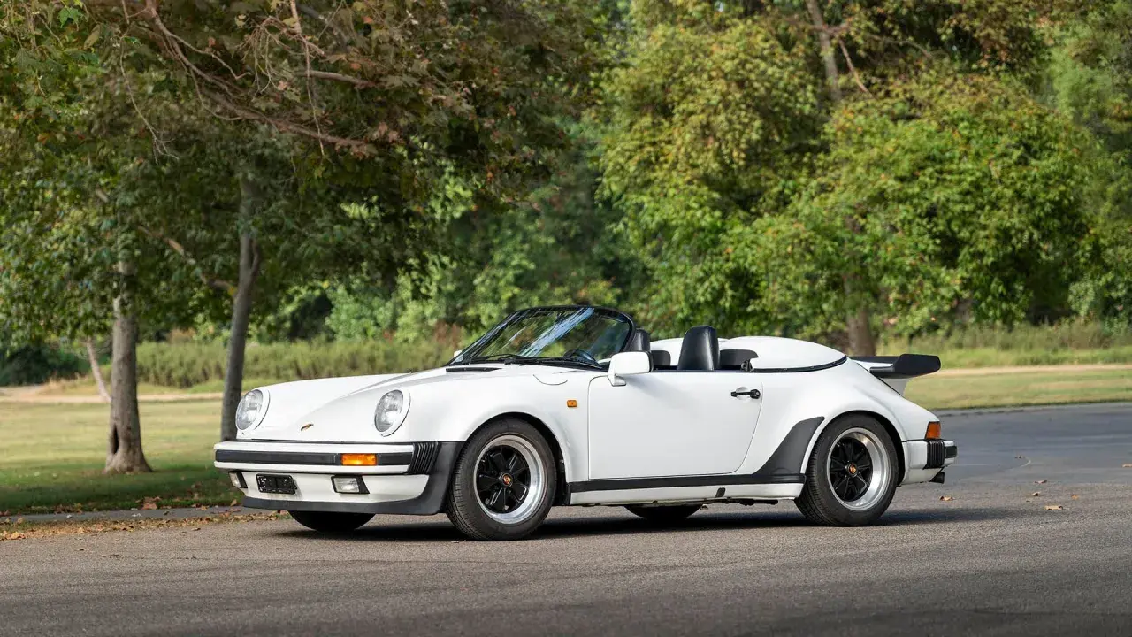 Biały Porsche 911 Speedster z lat 80. z czarnym dachem i spojlerem, zaparkowany na tle drzew.