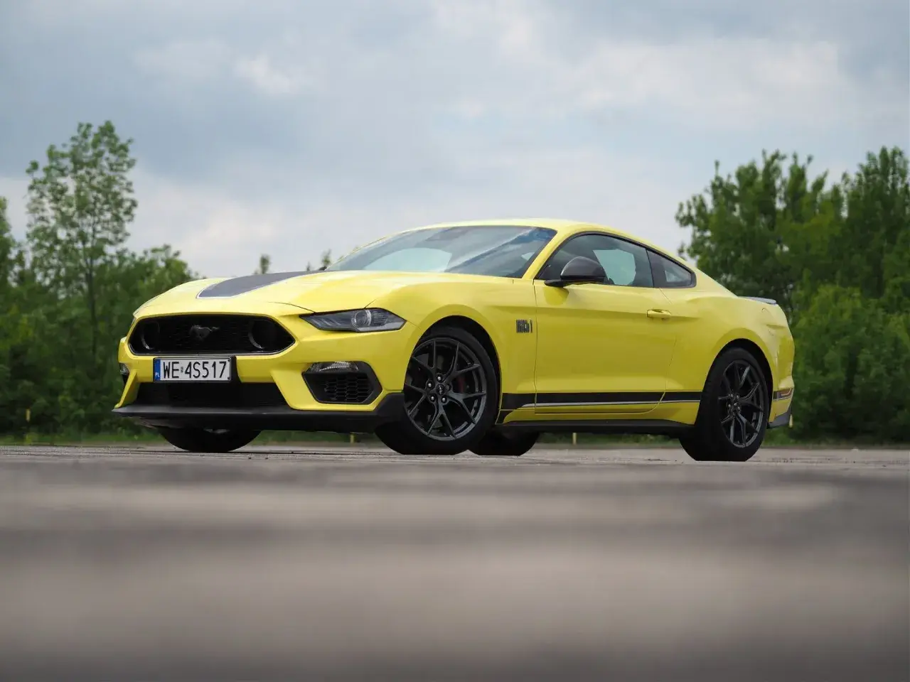 Żółty Ford Mustang Mach 1 z czarnymi pasami na masce i bokach, zaparkowany na tle zieleni.
