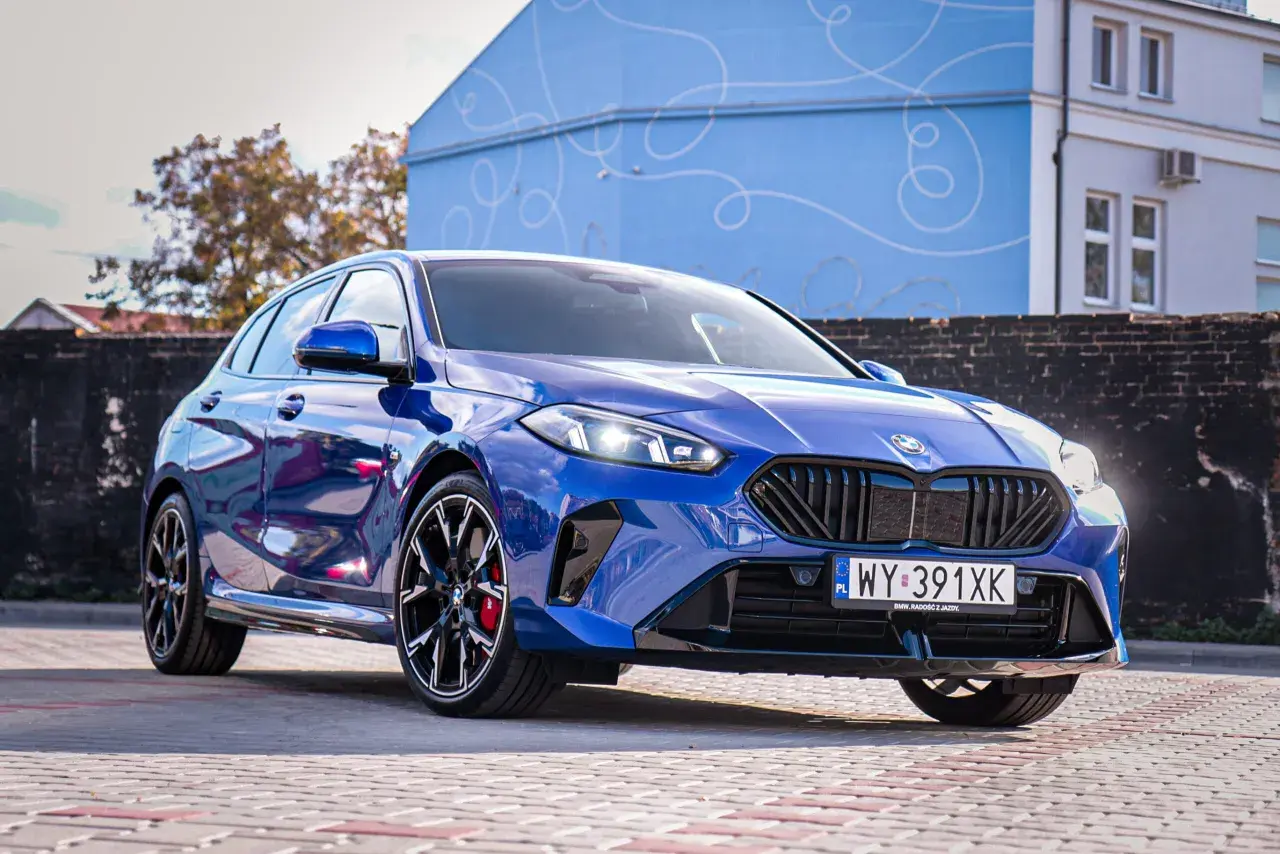 Błyszczące, niebieskie BMW 120 z czarnymi felgami stoi na brukowanym placu przed budynkiem z niebieską, ozdobną fasadą.