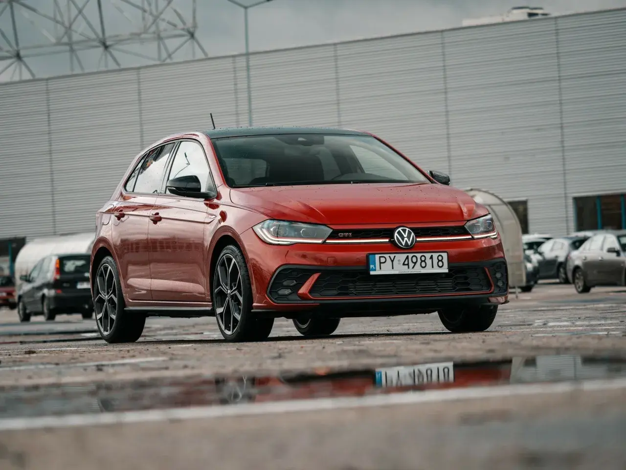 Czerwony Volkswagen Polo GTI z czarnym dachem, zaparkowany na mokrym parkingu.