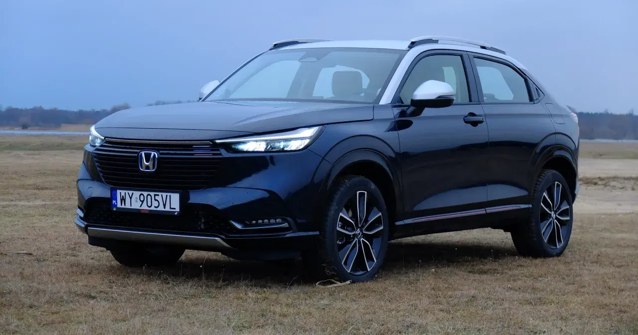 Nowa Honda HR-V w ciemnym kolorze, zaparkowana na trawiastym polu, z widokiem na jezioro i las w tle.