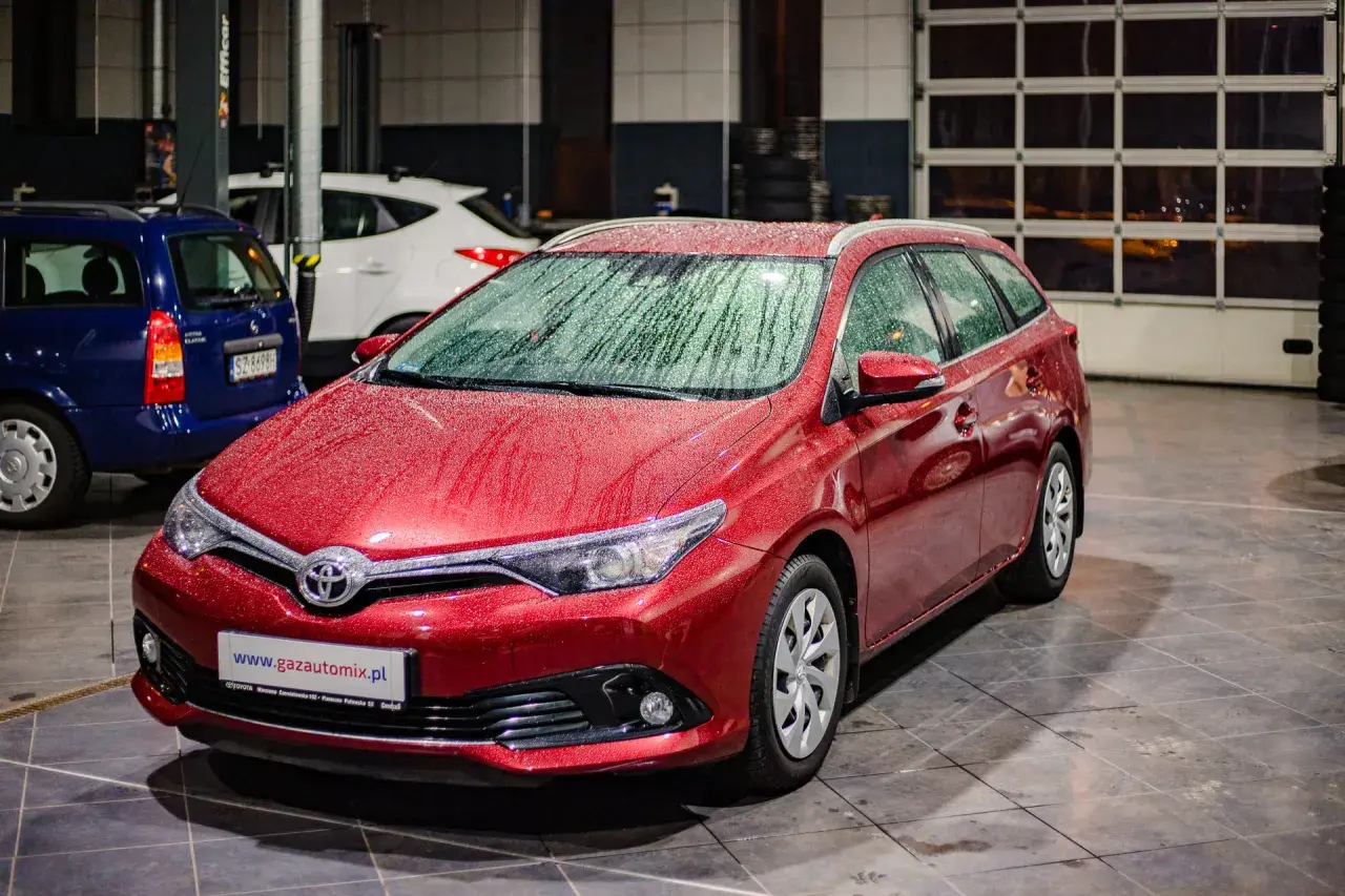 Czerwona Toyota Auris 1.6 benzyna z kroplami deszczu na karoserii, zaparkowana w garażu obok innych samochodów.