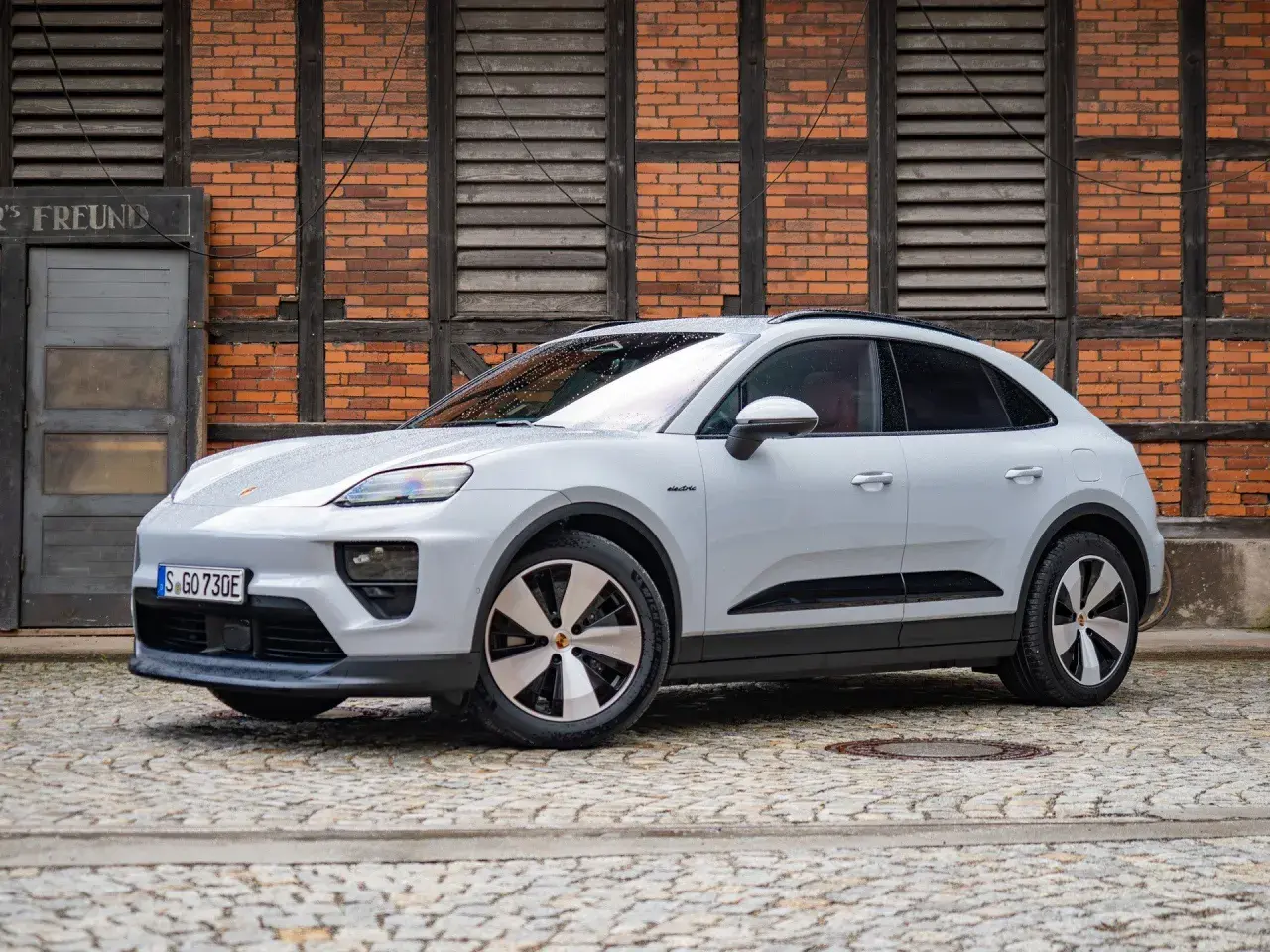 Biały Porsche Macan Electric na bruku, z ceglaną ścianą w tle. Sprawdź aktualną porsche macan cena.
