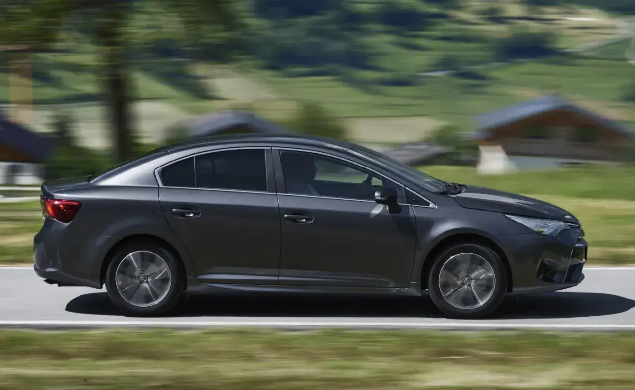 Ciemnoszara Toyota Avensis 2.0 D4D w ruchu, z rozmytym tłem górskiego krajobrazu.