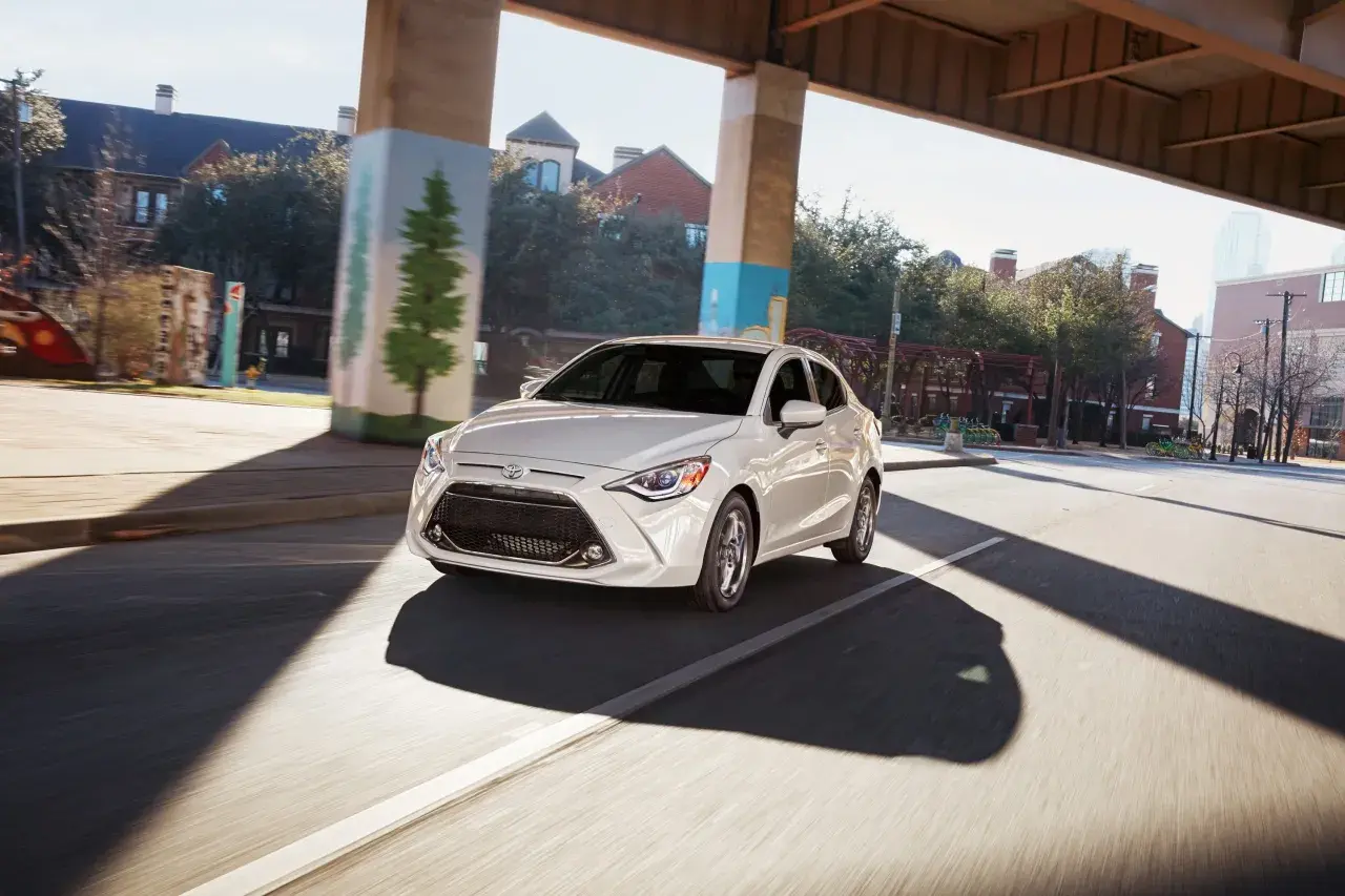 Biały Toyota Yaris Sedan jedzie pod wiaduktem, mijając budynki i drzewa.