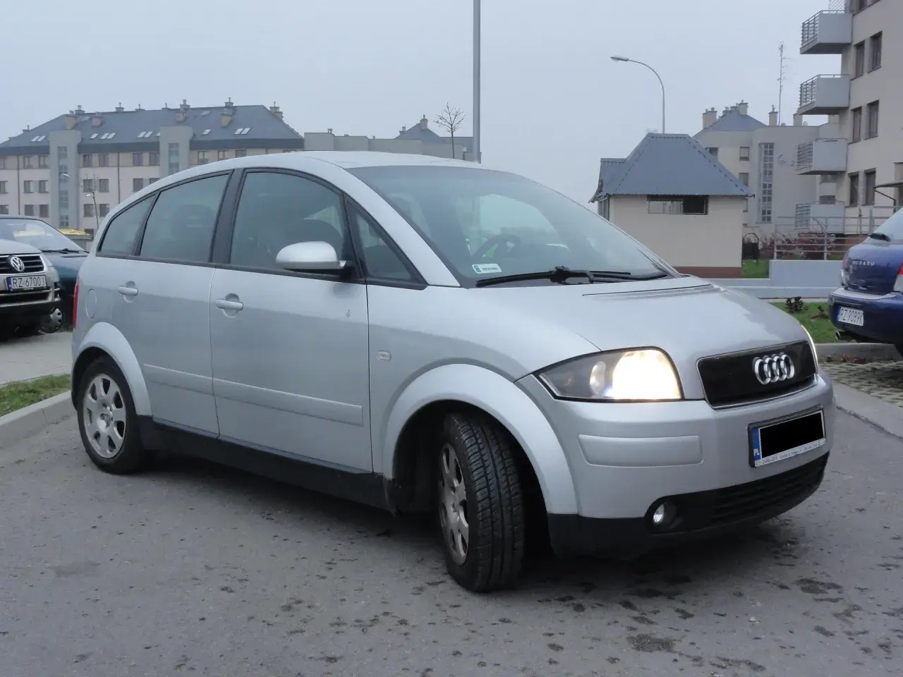 Srebrny Audi A2 1.4 benzyna zaparkowany przed blokami.