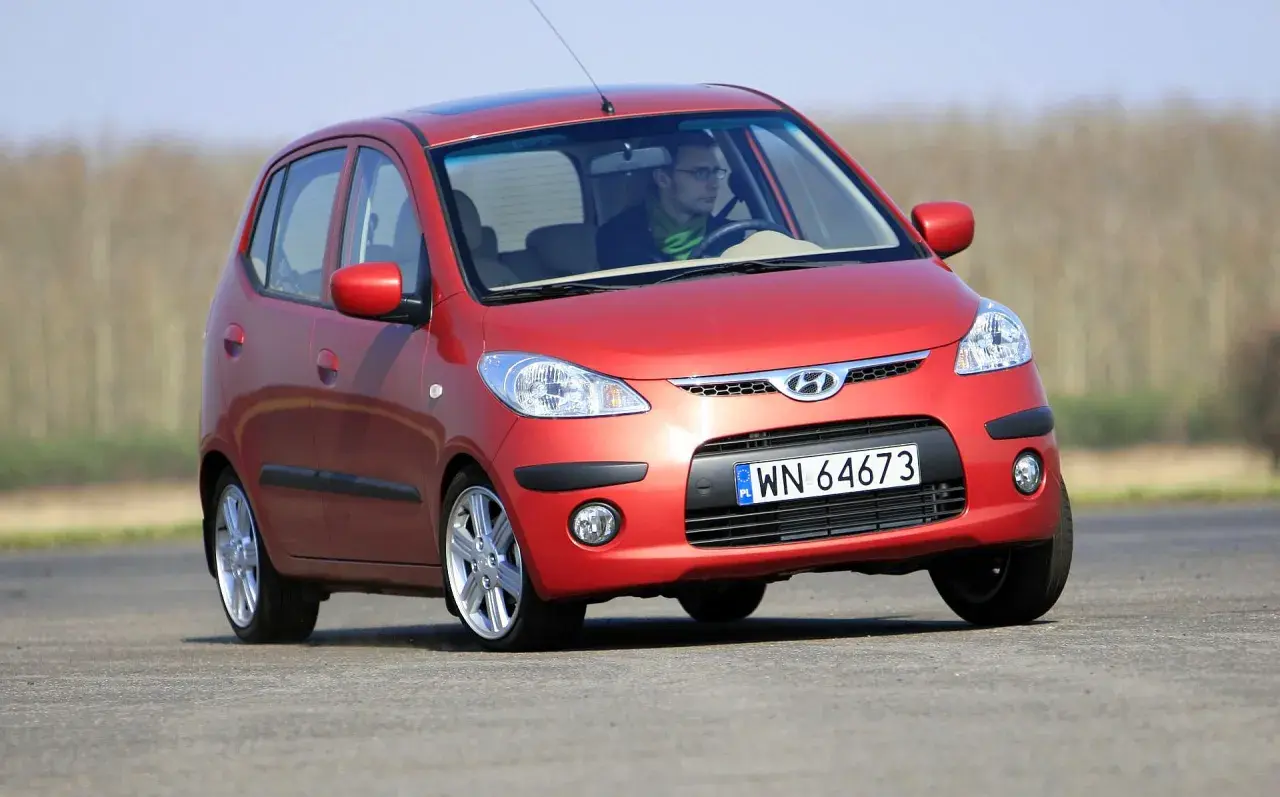 Czerwony Hyundai i10 na drodze. Kierowca w okularach i zielonym szaliku. Wiele pozytywnych hyundai i10 opinie potwierdza jego miejski charakter.