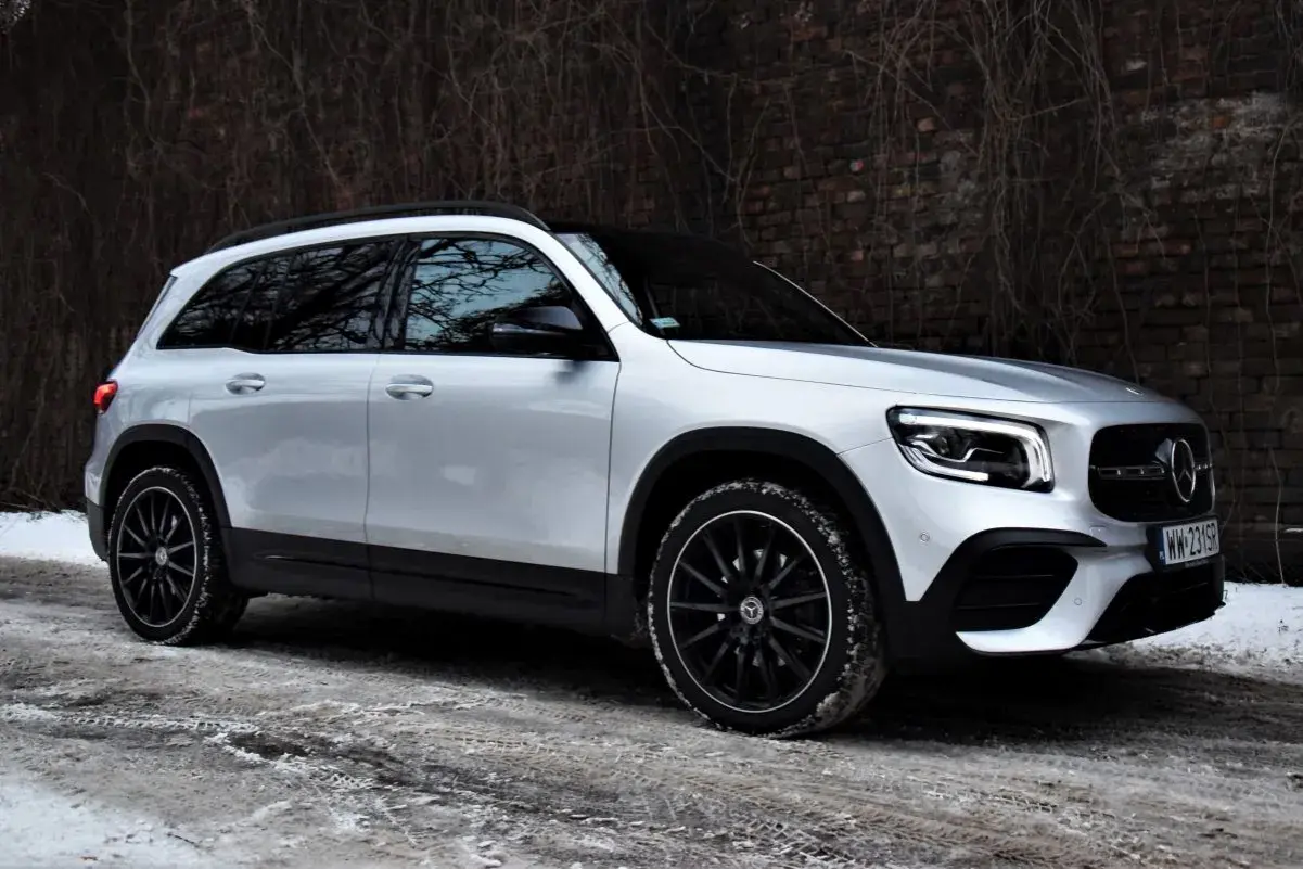 Srebrny Mercedes GLB 200 na czarnych felgach, zaparkowany na zaśnieżonej drodze przy ceglanym murze.