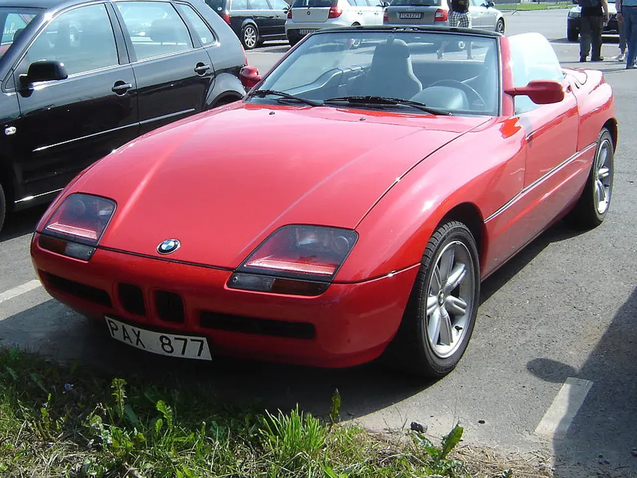 Czerwony kabriolet BMW Z1 z tablicą rejestracyjną PAX 877 zaparkowany obok czarnego samochodu.