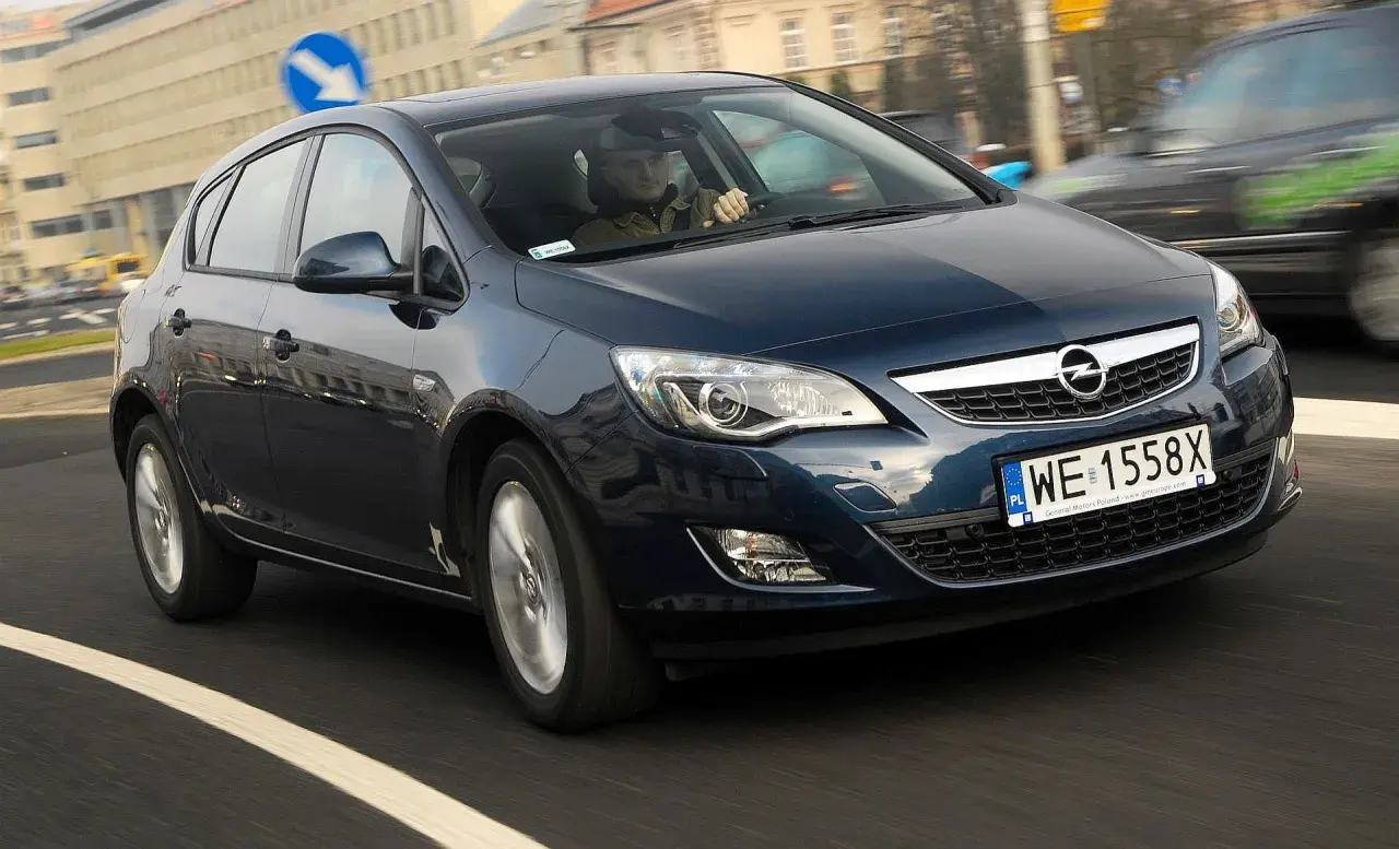 Ciemnoniebieski Opel Astra J 1.7 CDTI porusza się po miejskiej drodze. Kierowca w czapce prowadzi auto.