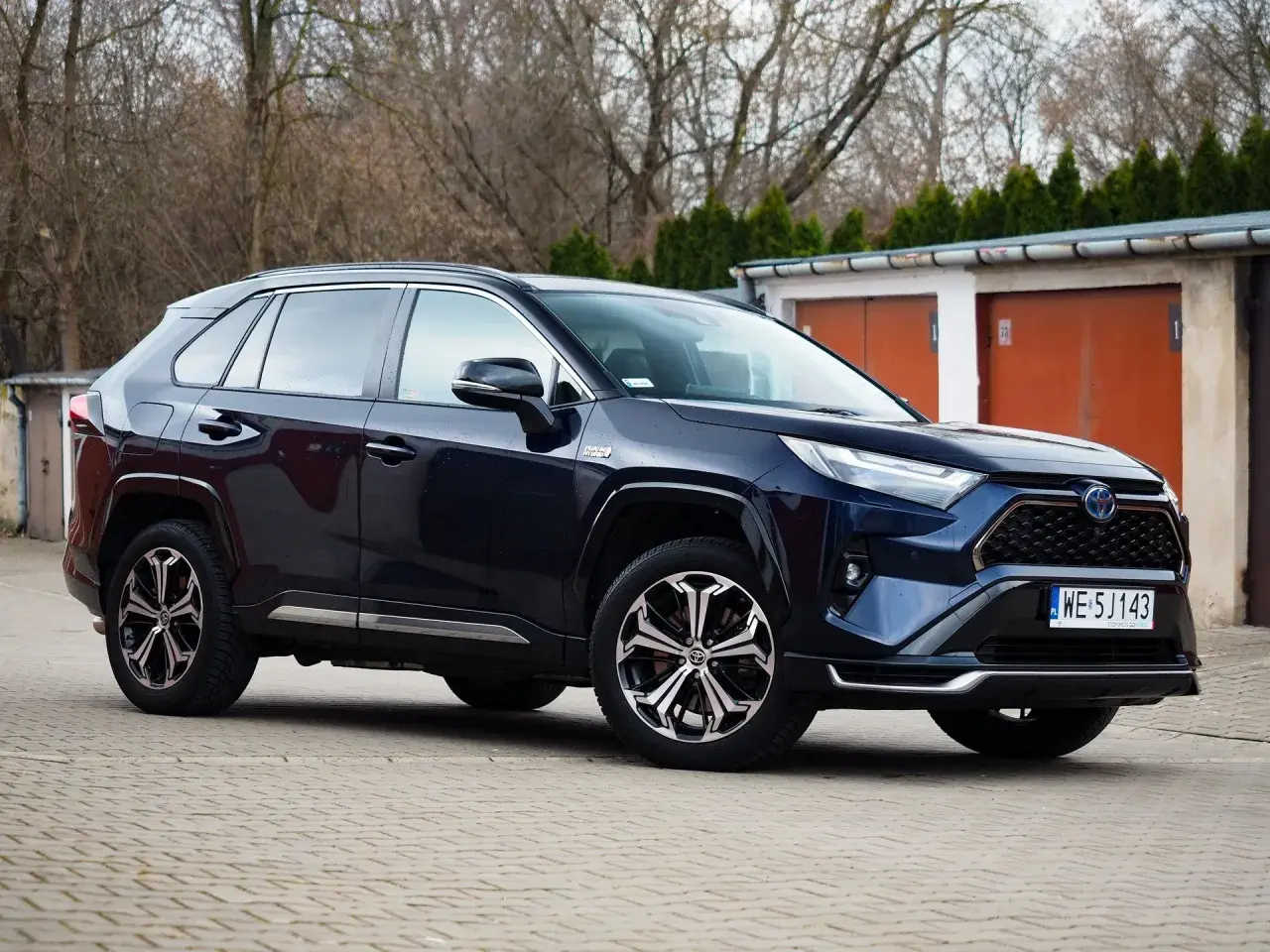 Ciemnoniebieski Toyota RAV4 Hybrid, często chwalony w toyota rav4 opinie, zaparkowany przed garażami.