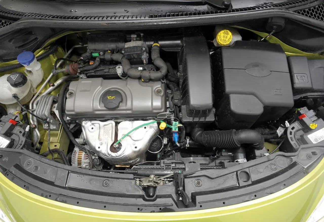 Silnik peugeot 207 1.4 benzyna. Widok komory silnika z widocznymi elementami, takimi jak alternator, obudowa filtra powietrza i zbiorniczek płynu chłodniczego.