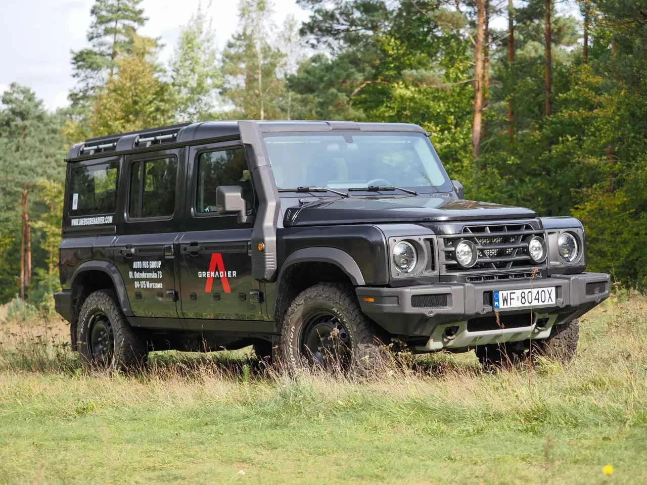 Czarny grenadier auto z bagażnikiem dachowym i snorkelami, zaparkowany na trawie wśród drzew.