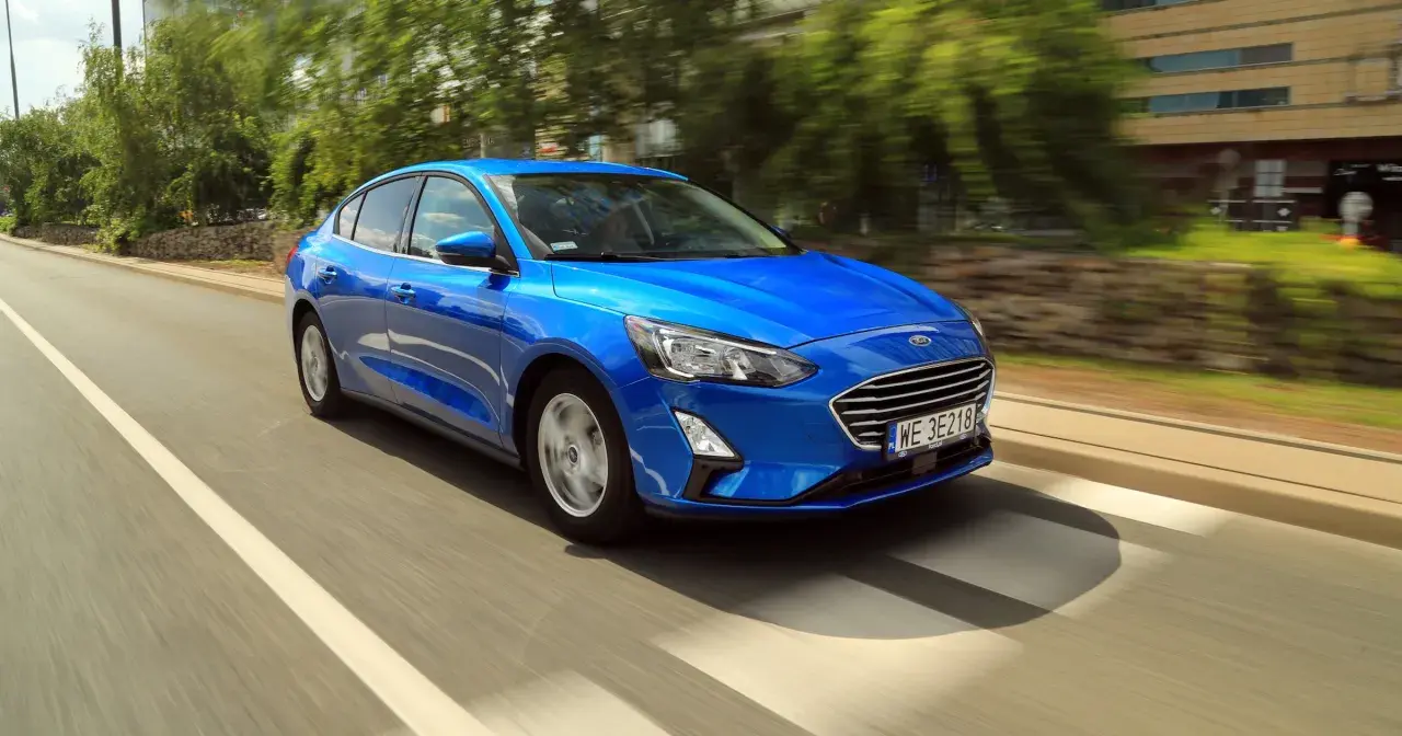 Błękitny Ford Focus sedan pędzi po miejskiej drodze, mijając drzewa i budynki.