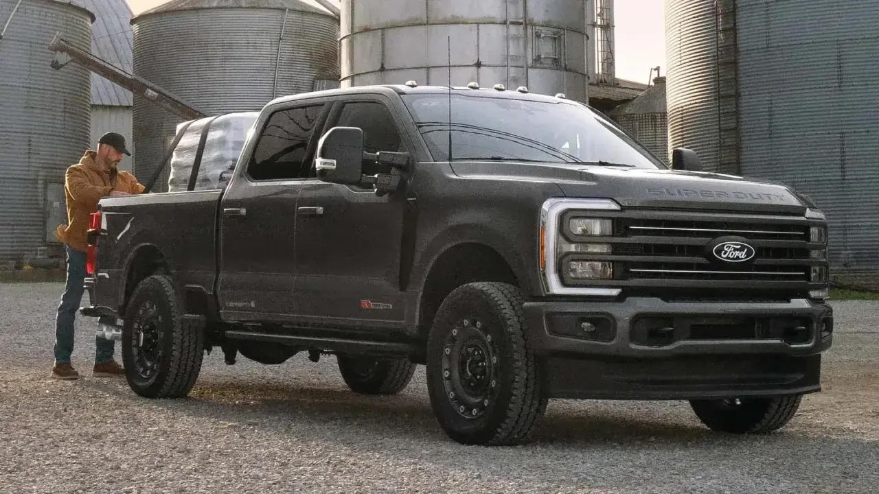 Mężczyzna ładuje pakunki do ciemnoszarego Forda Super Duty. Ciężarówka stoi na żwirowej drodze obok silosów.