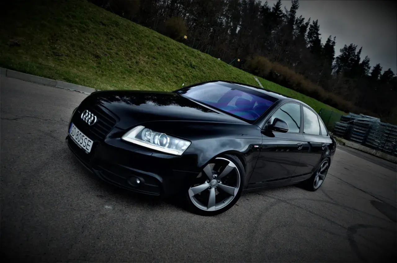 Czarny Audi A6 C6 3.0 TDI Quattro S-line na tle zielonego wzgórza i lasu.