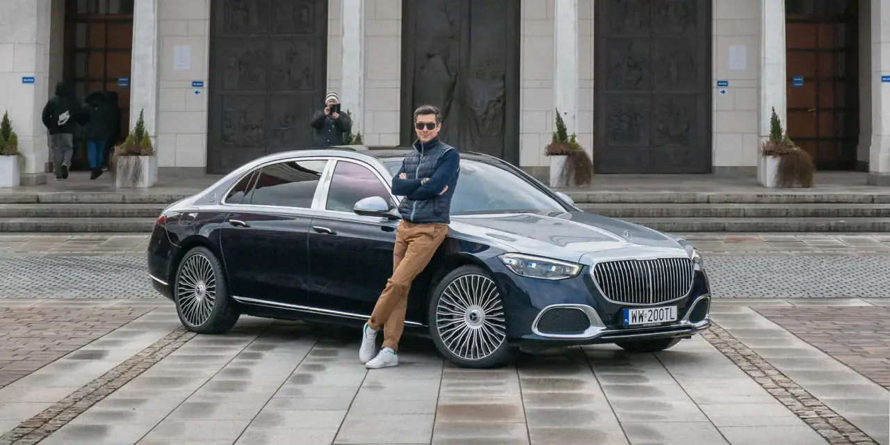 Elegancki mężczyzna opiera się o lśniącego Maybacha S580 przed monumentalnym budynkiem.