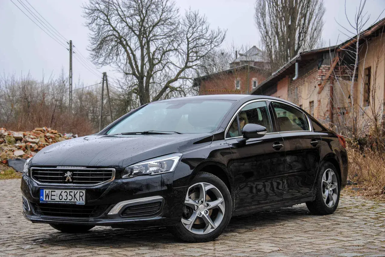 Czarny Peugeot 508 na brukowanej drodze. W tle zrujnowane budynki i drzewa. Wiele osób szuka peugeot 508 opinie przed zakupem.