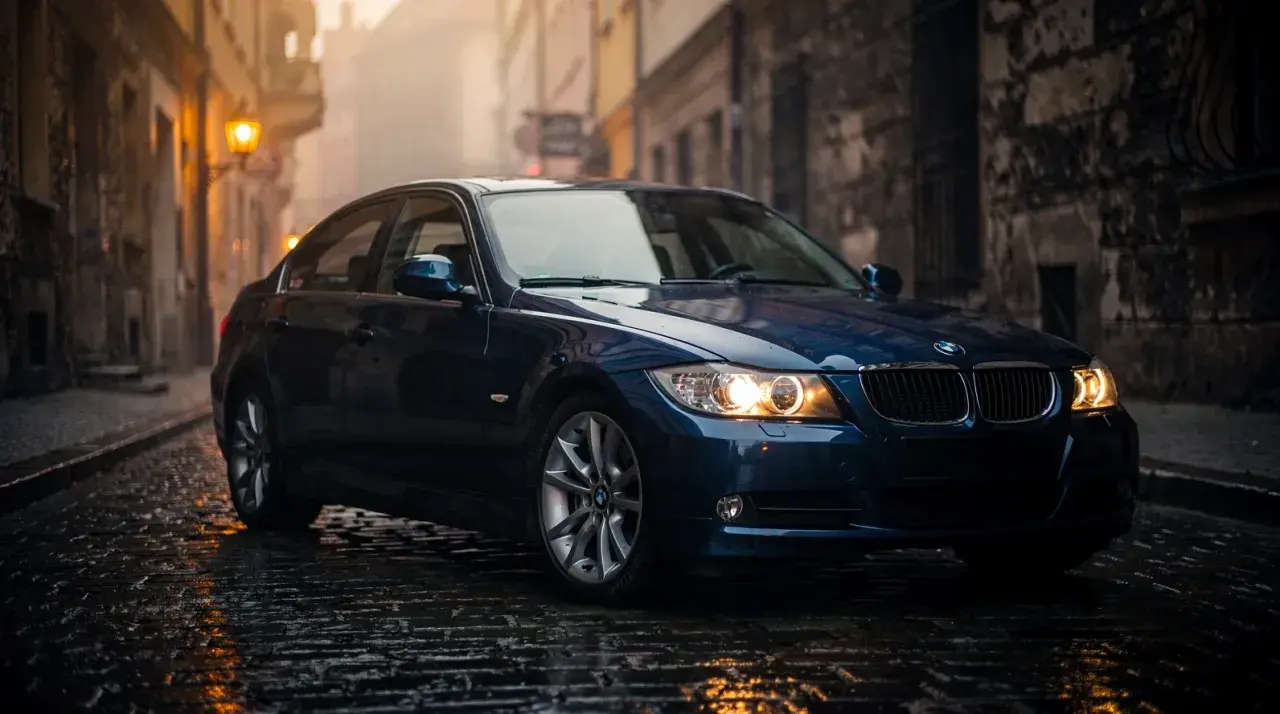 Ciemnoniebieskie BMW E90 2.0 benzyna na mokrej, brukowanej uliczce w starym mieście.