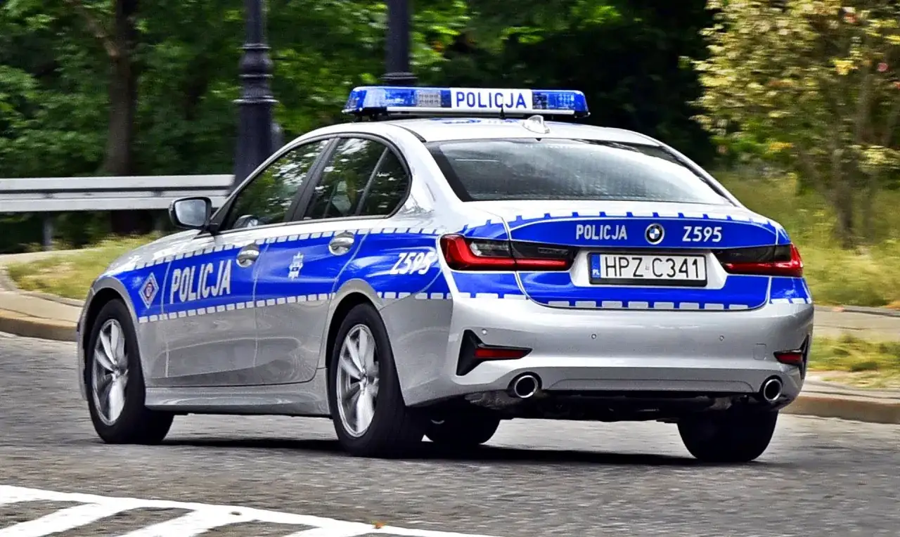 Srebrny radiowóz policyjny z niebieskimi pasami i napisem "POLICJA" na drzwiach i tyle. Widoczna tablica rejestracyjna HPZ C341.
