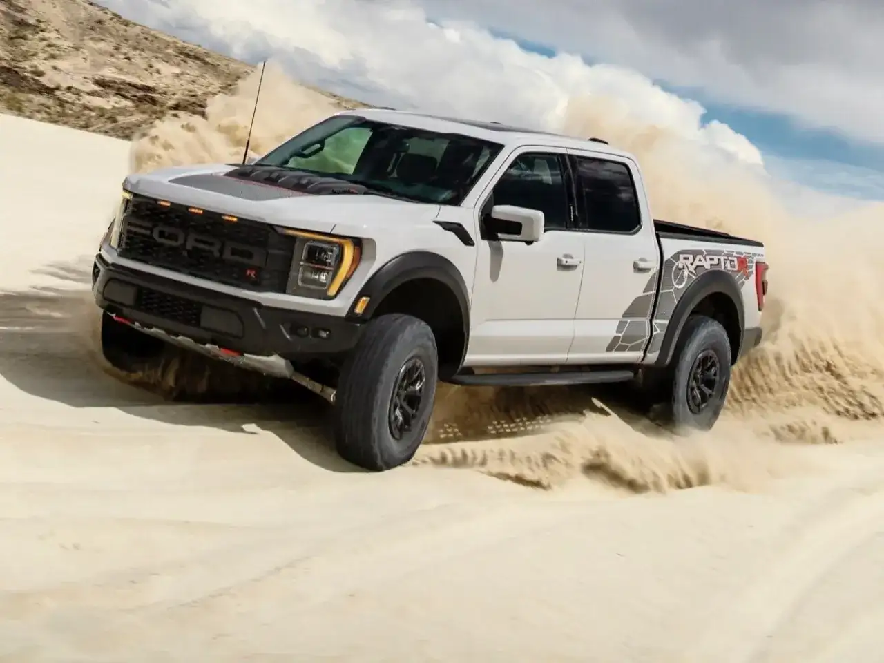 Biały Ford F-150 Raptor szaleje po piaszczystych wydmach, wzbijając tumany piasku.