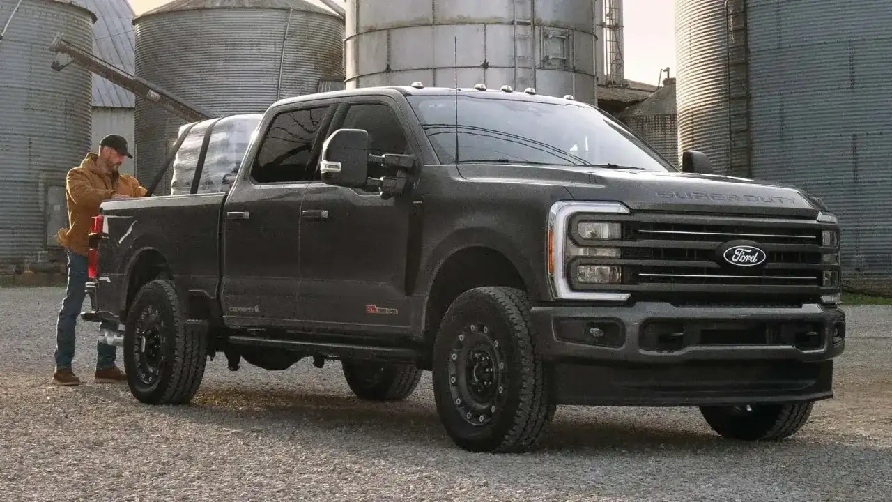 Mężczyzna ładuje pakunki do ciemnoszarego Forda Super Duty. Ciężarówka stoi na żwirowej drodze obok silosów.