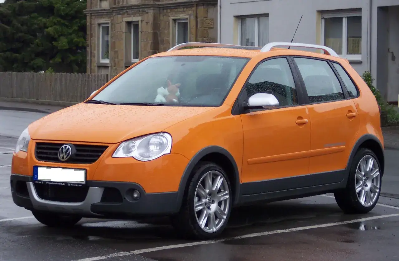 Pomarańczowy VW Cross Polo na mokrym parkingu, z pluszowym misiem za przednią szybą.