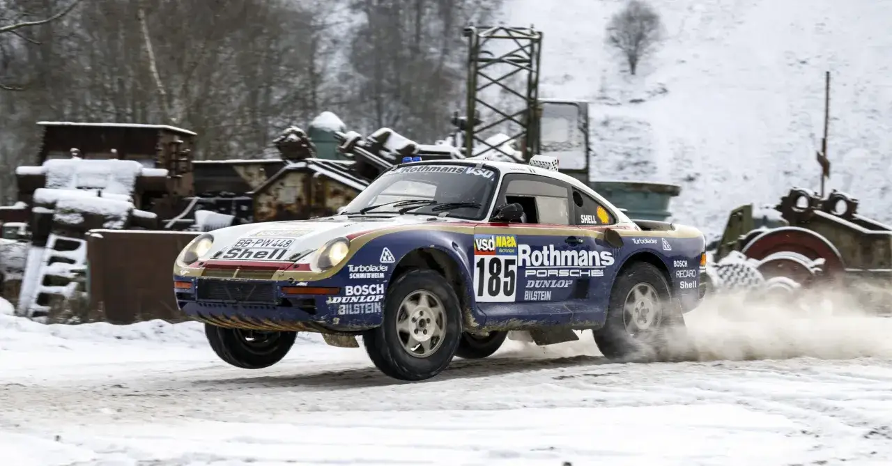 Porsche 959 w barwach Rothmans, z numerem 185, pokonuje śnieżny zakręt, wzbijając tumany śniegu.