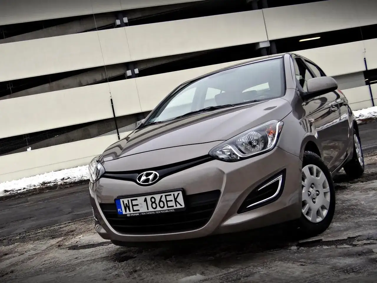 Brązowy Hyundai i20 1.2 benzyna zaparkowany przed nowoczesnym budynkiem.