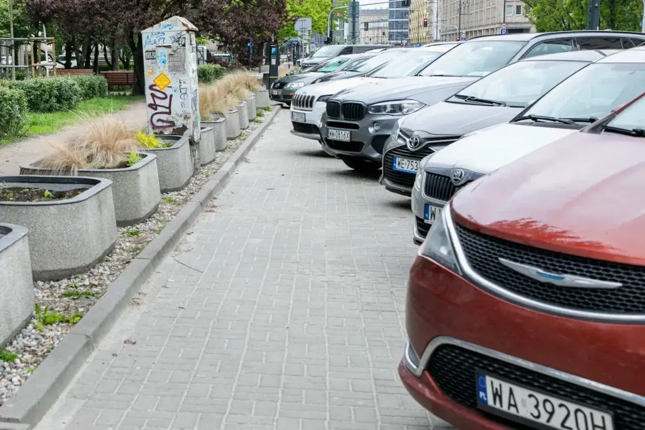 Rząd samochodów zaparkowanych na chodniku, blokujących przejście. Betonowe donice z trawą ozdobną tworzą barierę.