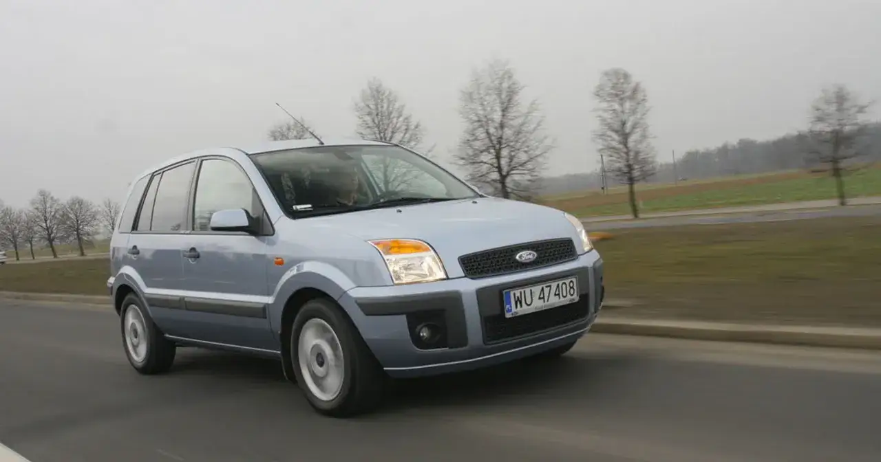 Srebrny Ford Fusion 1.4 benzyna jedzie po drodze, za nim rozciąga się krajobraz z drzewami i polami.