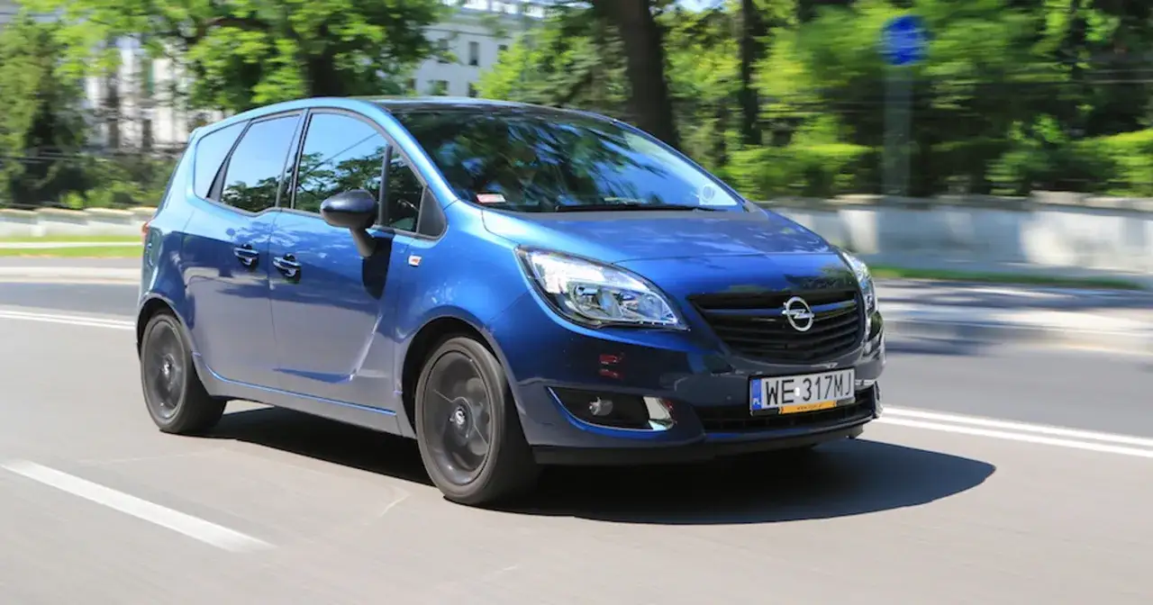 Niebieski Opel Meriva 1.6 benzyna w ruchu miejskim, z rozmytym tłem drzew i budynków.