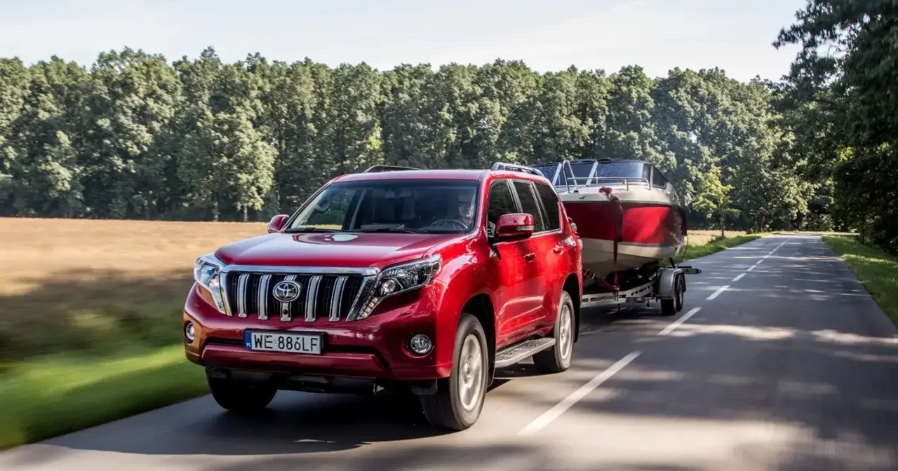 Czerwony RAM 2500 holuje łódź motorową po wiejskiej drodze wśród drzew.